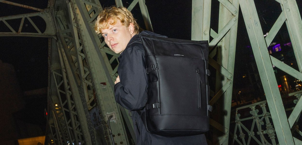 Černý batoh s nastavitelnými popruhy, přední kapsou na zip a hladkým povrchem. Nosený na zádech na nočním pozadí.