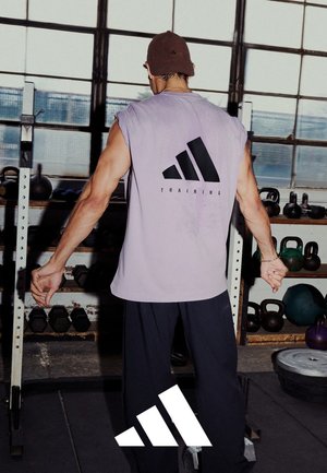 Lila ärmelloses Shirt mit schwarzem Branding auf dem Rücken, kombiniert mit dunklen Hosen, in einer Fitnessumgebung mit sichtbaren Gewichten.
