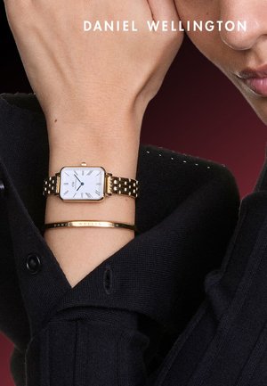 Montre-bracelet en or avec un cadran rectangulaire et un fond blanc, associée à un bracelet assorti en or avec du texte gravé. Portée au poignet, sur un fond en tissu noir.