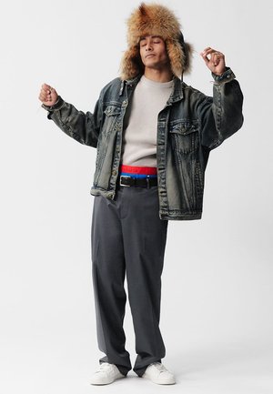 Jeansjacke mit pelzgefütterter Kapuze, hellgrauer Pullover, graue Hose, roter und blauer Gürtel, weiße Turnschuhe, stehende Pose vor einem einfarbigen Hintergrund.
