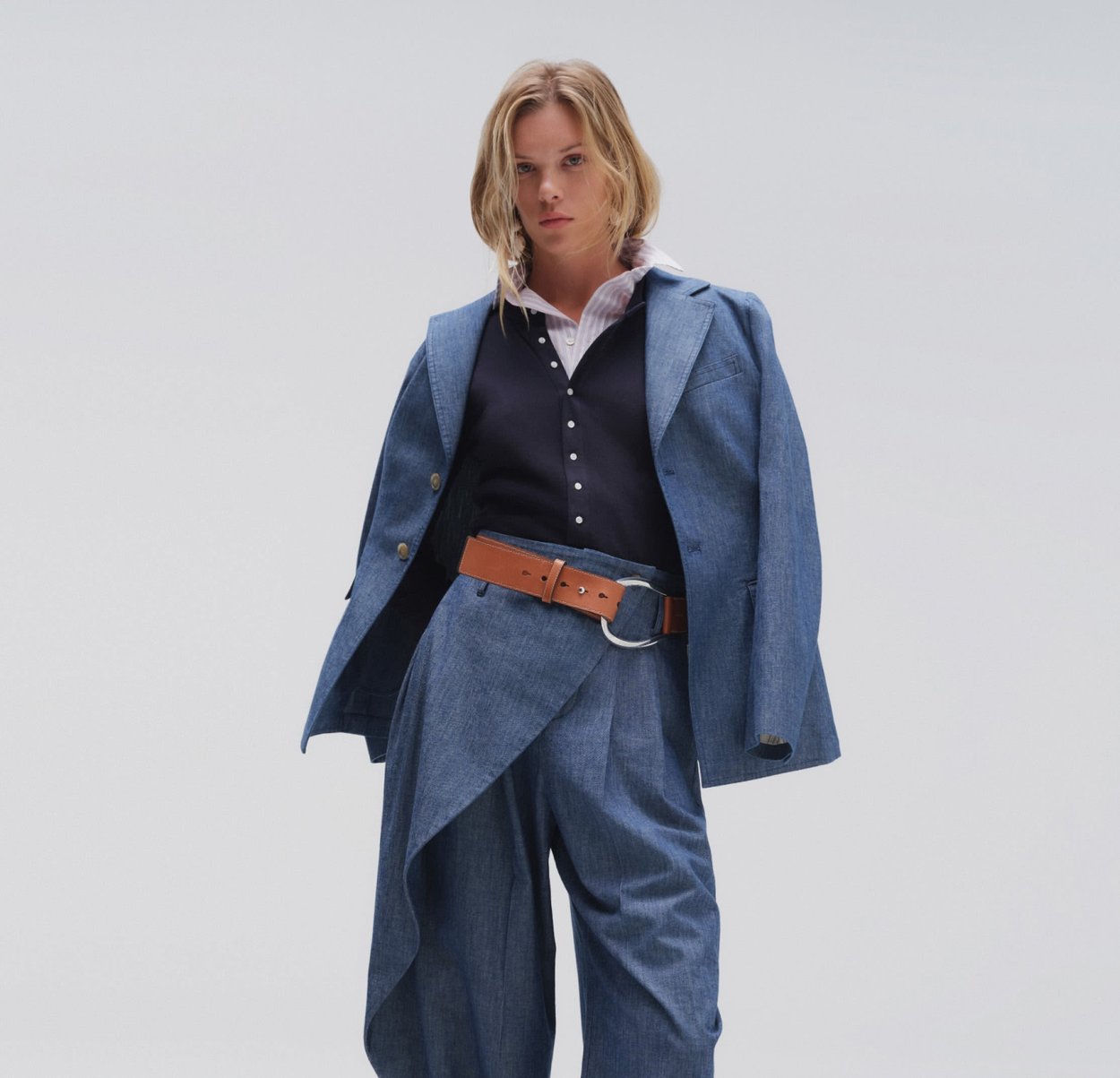 Mujer con un traje azul oversize con pantalones de pierna ancha, un cinturón marrón, chaleco negro y camisa blanca con cuello, sobre un fondo liso.
