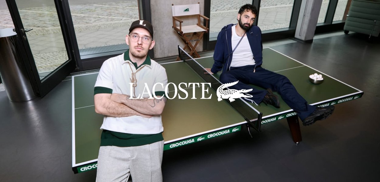 Zwei Männer posieren an einem grünen Crocoulga-Tischtennistisch in einem hellen Raum mit großen Fenstern und einem Regiestuhl.