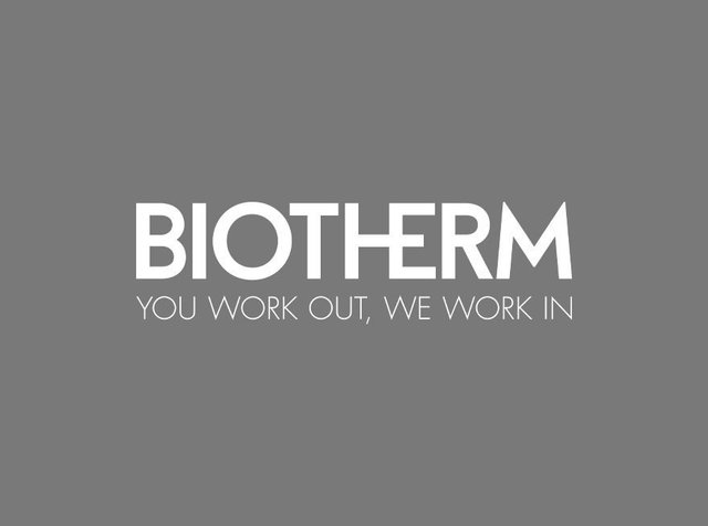 Weisser Text "BIOTHERM" über dem Satz "DU TRAINIERST, WIR ARBEITEN" auf grauem Hintergrund. Die Schrift ist fett und modern mit klarer Lesbarkeit.