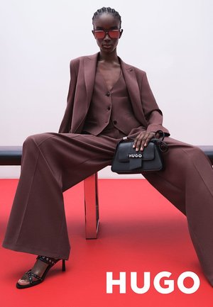Completo tailored bordeaux con pantaloni larghi e gilet aderente, abbinato a una borsa nera con logo e tacchi con cinturini. Sfondo rosso.