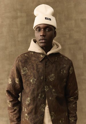 Ung mand iført en cremefarvet hue, beige hættetrøje og brun jakke med blomsterprint, stående mod en ensfarvet beige baggrund.