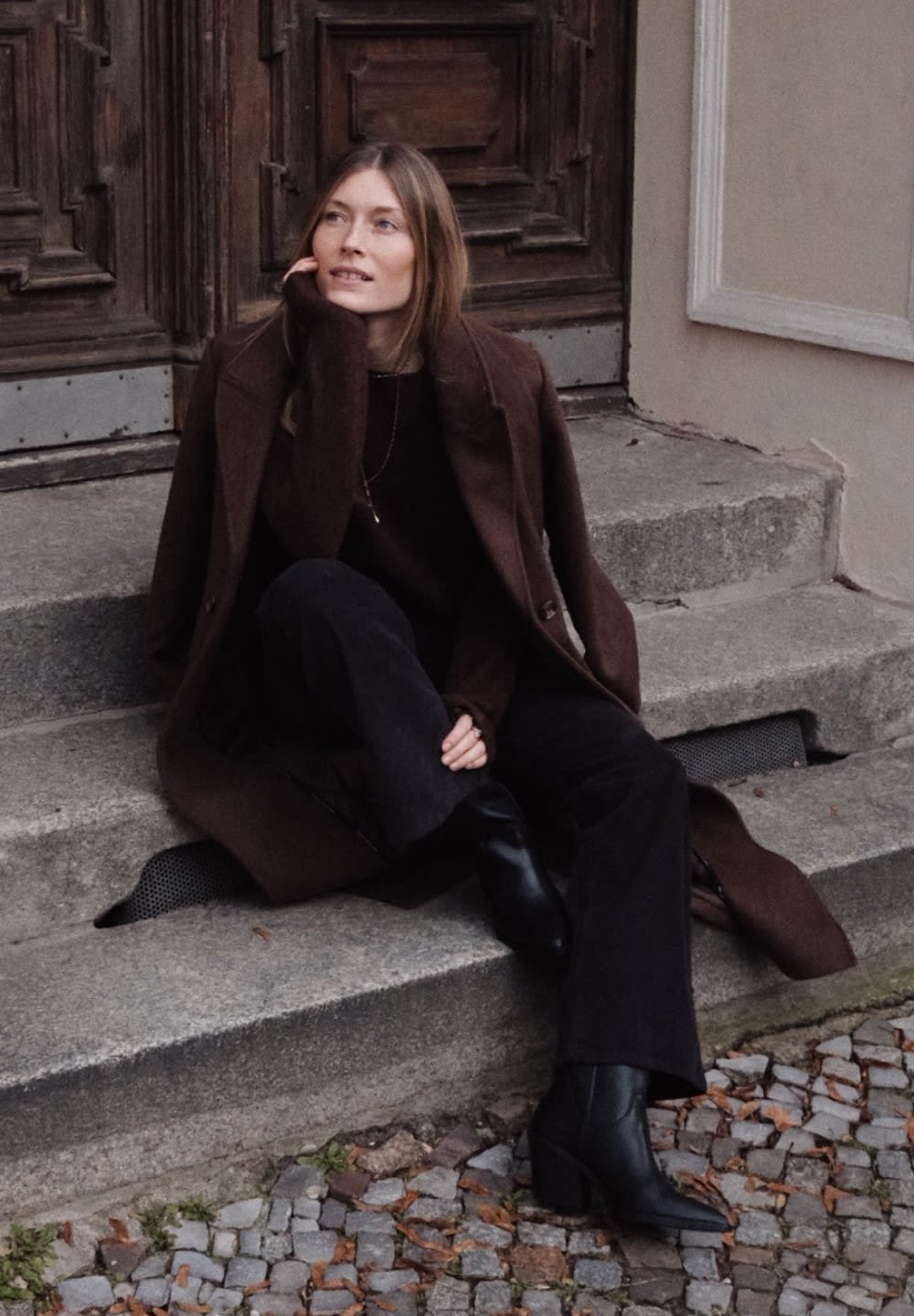 Abrigo de lana marrón sobre un suéter negro y pantalones acampanados negros, combinado con botines negros de tacón, sentado en escalones de piedra.