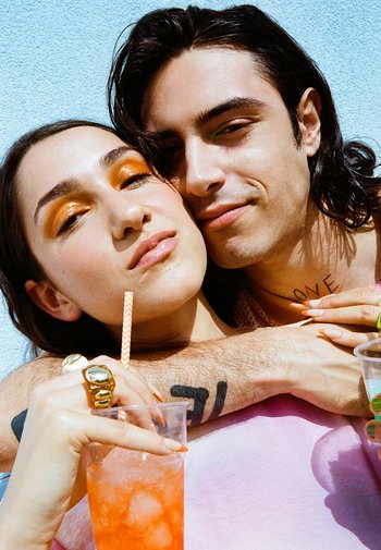 Due persone si abbracciano, entrambe con drink colorati. La persona davanti ha ombretto arancione, indossa anelli con grandi pietre e una maglietta rosa.