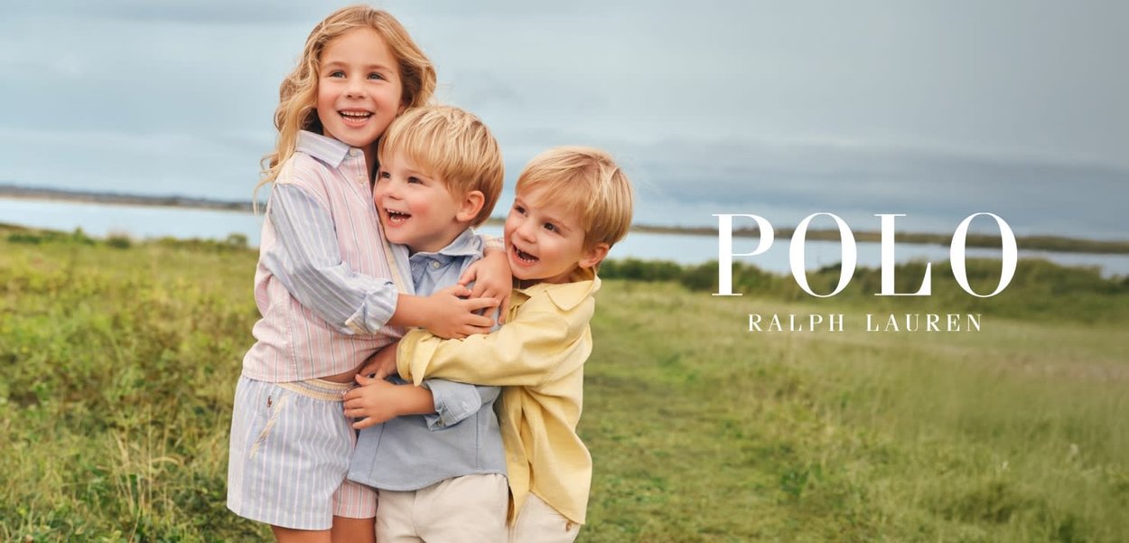 Tres niños pequeños sonriendo y abrazándose al aire libre cerca del agua, vestidos con ropa de colores pastel de Ralph Lauren.