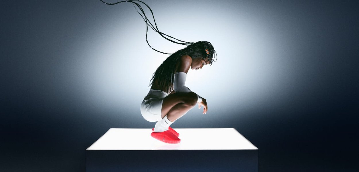 Calzature rosse, abbigliamento atletico bianco, figura accovacciata con lunghi capelli intrecciati, illuminata da una superficie bianca luminosa su uno sfondo scuro.