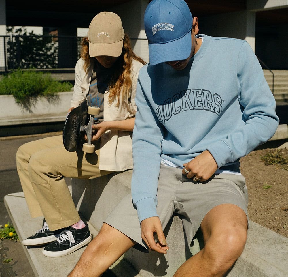 Un sweat-shirt bleu clair avec le texte "DOCKERS", un short gris et des baskets. Une femme portant une casquette beige tient un skateboard noir, vêtue de pantalons kaki.