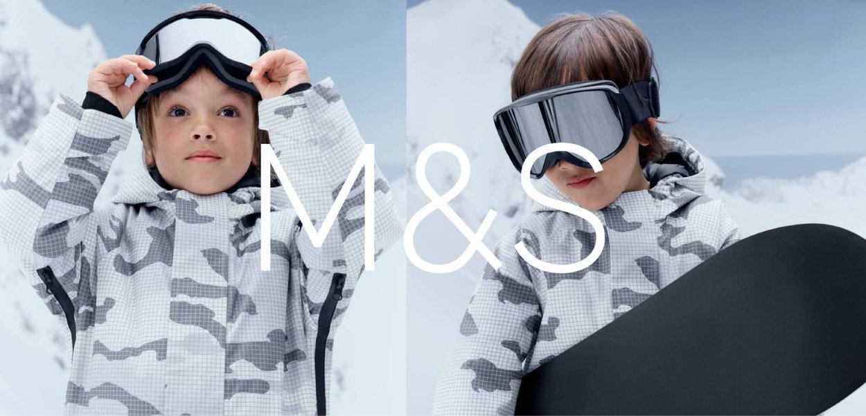 Enfant en veste de ski camouflage blanc-gris ajustant ses lunettes de ski, tenant un snowboard, avec un paysage montagneux enneigé en arrière-plan.