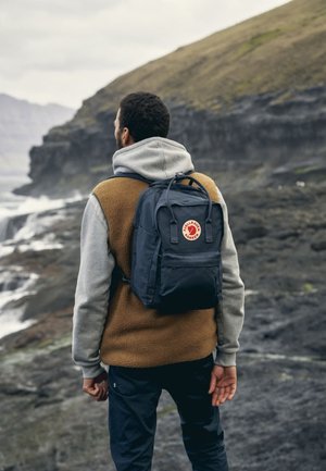 Marineblauer Rucksack mit einem roten Logo, aus Stoffmaterial, rechteckige Form, verstellbare Träger, getragen von einer Person mit einem grau-braunen Mantel.