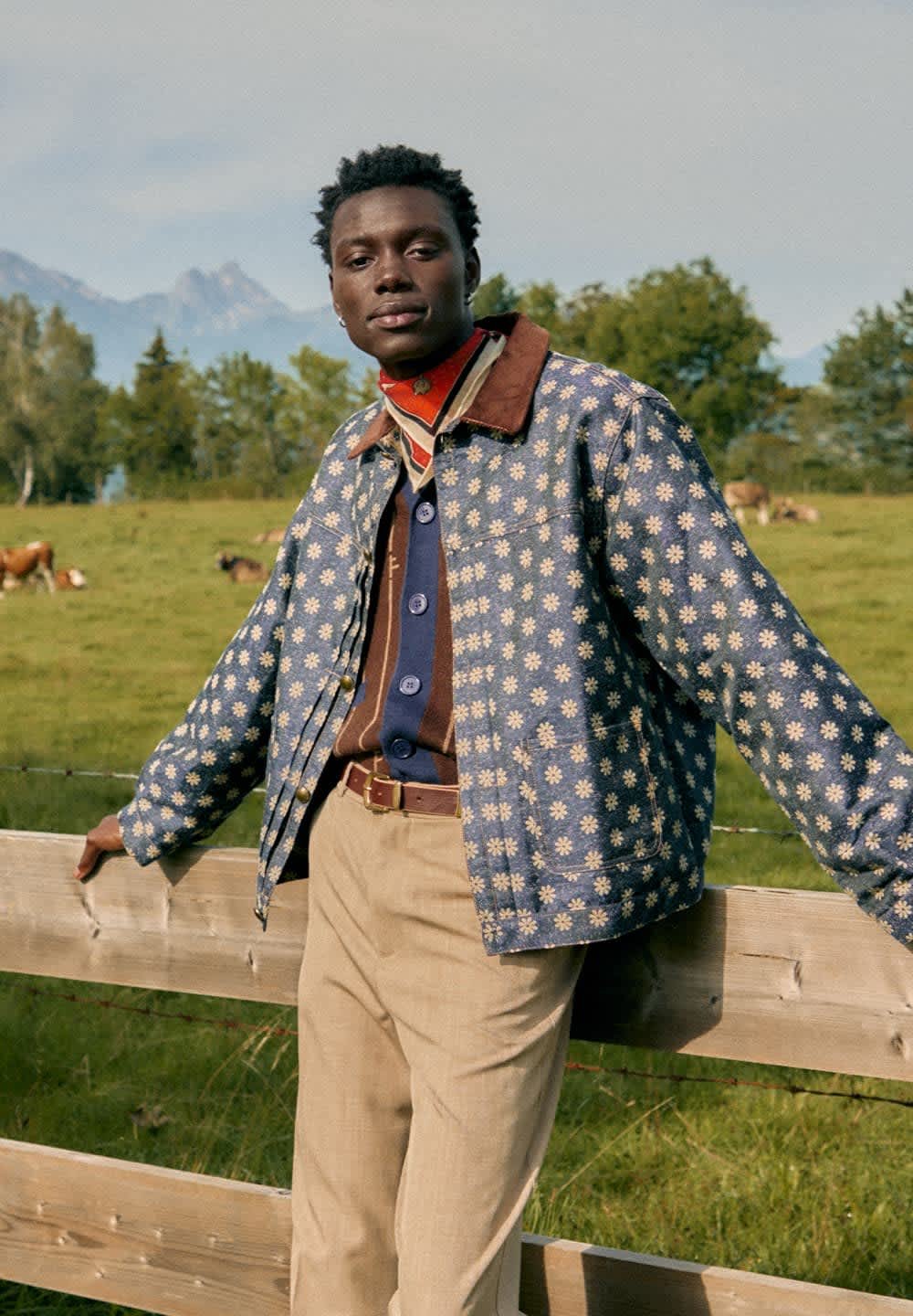 Veste en denim à motif floral avec un col marron, boutons bleus, superposée sur une chemise marron. Un pantalon beige et une écharpe rouge complètent le look.