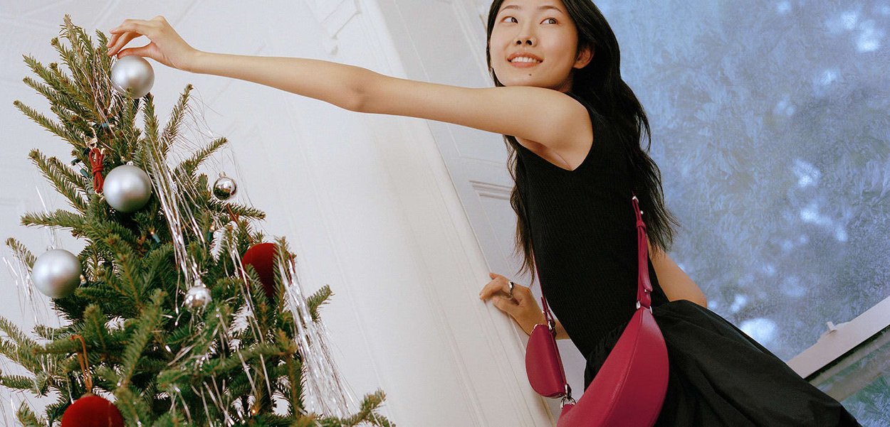 Borsa magenta con design curvo, indossata a tracolla. Albero di Natale decorato con ornamenti argento e rosso, fili argentati e fogliame verde.