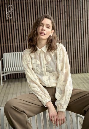 Frau mit welligem braunem Haar, die eine durchsichtige Bluse mit Blumenmuster und braune Hosen trägt, sitzt auf einem weißen Stuhl vor einer Wand aus hölzernen Latten.