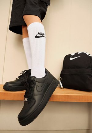 Beine in schwarzen Shorts, weißen Nike Socken und schwarzen Nike Air Force 1 Sneakers stehen auf einer Holzbank, neben ihnen ein schwarzer Nike Rucksack.