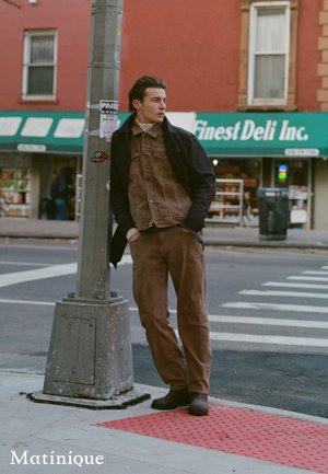 Giovane uomo con giacca e pantaloni marroni appoggiato a un lampione su un marciapiede della città vicino a una gastronomia e a una strisce pedonali.