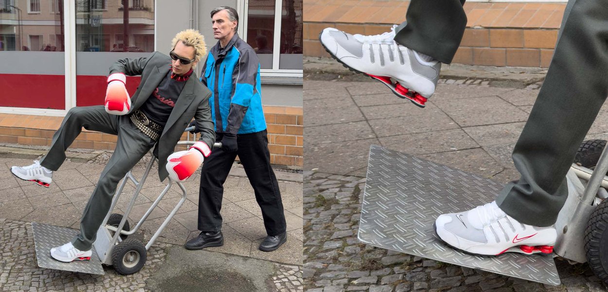 To mænd i urbant miljø. Den ene bærer en grå jakkesæt med et bælte med nitter og store røde boksehandsker, mens den anden skubber en håndvogn.