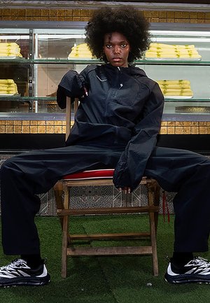Black lightweight jacket and pants, featuring a zip closure, relaxed fit, paired with black and white athletic shoes. Sitting on a wooden chair.