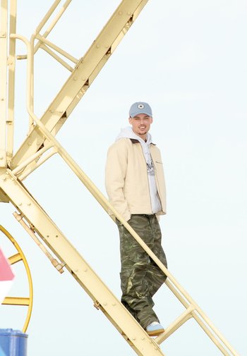 Beige jas over een grijze hoodie, camouflagebroek en lichtblauwe sneakers. Staande op een gele metalen trap met een heldere achtergrond.