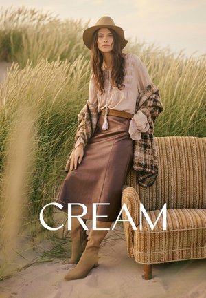 Brown leather skirt, cream blouse with embroidery, grey patterned shawl, tan hat, and knee-high beige boots, against grass backdrop.