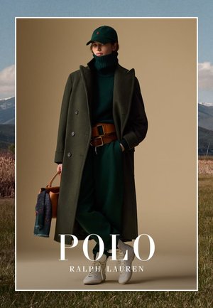 Personne portant un manteau vert foncé, un pull col roulé, une casquette, une large ceinture, un pantalon vert, des baskets blanches, tenant un sac à main marron avec une écharpe bleue.