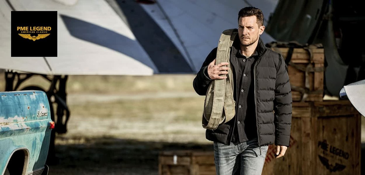 Veste matelassée noire et chemise sombre portées par un homme portant un sac en toile. En arrière-plan, un avion et des caisses en bois.