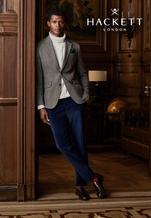 Blazer gris con estampado pata de gallo sobre un suéter de cuello alto color crema, pantalones azules y zapatos oscuros. Pañuelo en el bolsillo y detalles texturizados en el traje visibles. Fondo de madera.