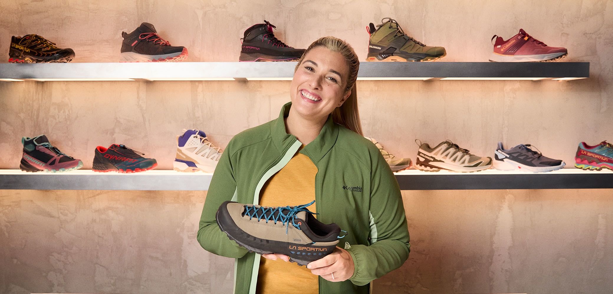 Hiking shoe in brown and black with blue laces and textured sole, held by a person wearing a green jacket, with various shoes displayed behind.