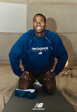 Giovane uomo con felpa blu New Balance inginocchiato su un tappetino grigio, sorridente e con lo sguardo rivolto verso l'alto, all'interno, con delle bande elastiche per esercizi accanto a lui.