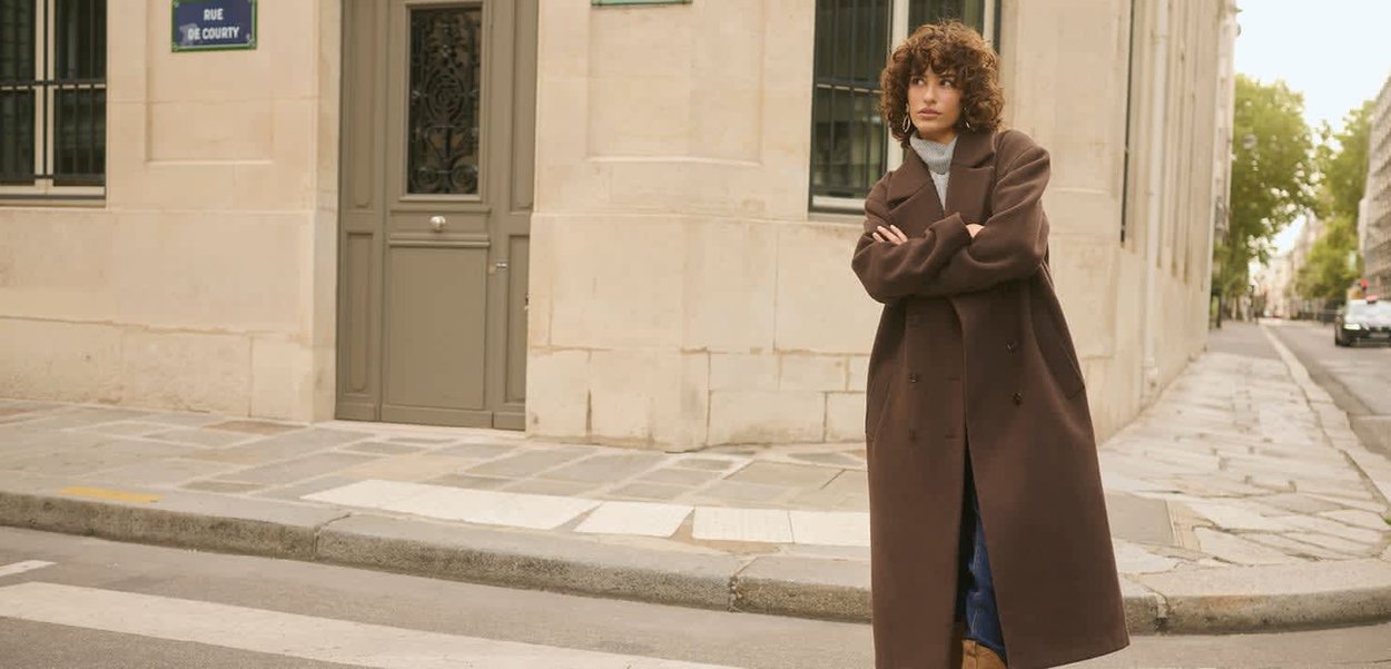 Ruskea ylisuurikokoinen takki lovikauluksella ja isoilla napeilla. Päällä harmaa poolopaita ja farkut, kaupungin kadulla kivenlaatoitetulla jalkakäytävällä.