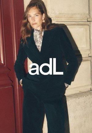 Costume texturé vert foncé avec une veste ajustée et un pantalon à jambes larges, associé à un chemisier floral clair et de grandes boucles d'oreilles en or.