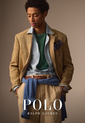 Blazer à chevrons beige sur une chemise bleue, un t-shirt vert et un pantalon beige, avec un pull bleu drapé et un pochette. Ceinture marron et montre.