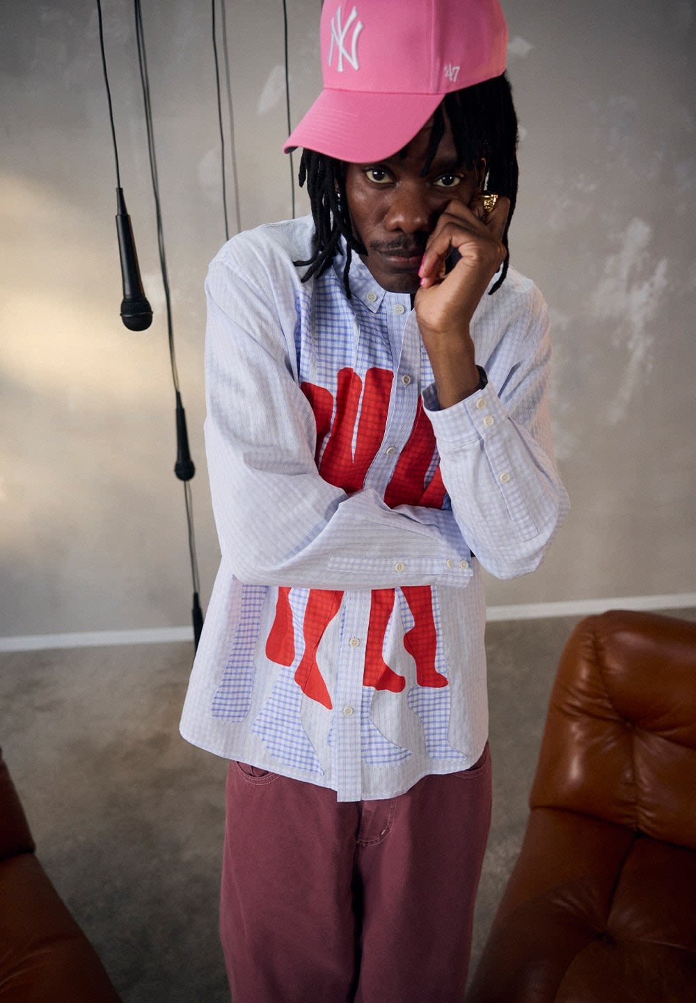 Camicia a maniche lunghe in gingham azzurro chiaro con un motivo grafico astratto rosso. Indossata con pantaloni color bordeaux e un cappellino rosa con un logo.