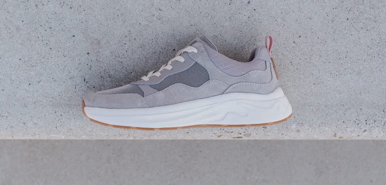 Grauer Sneaker aus Wildleder und Mesh, mit einer weißen Mittelsohle, Gummiaußensohle und pinken Akzenten an der Ferse. Glatte Textur und schlankes Design.