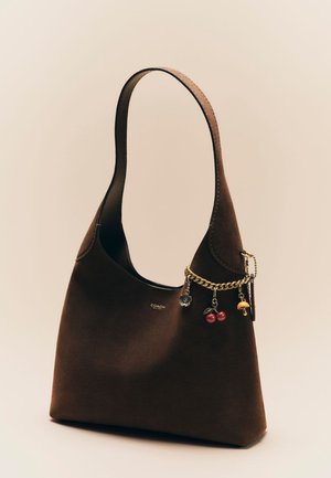 Braune Leder-Handtasche von Coach mit einem Tragegurt und einer goldenen Charmkette mit Anhängern in Form einer Blume, Kirschen und einem Pilz auf beigem Hintergrund.