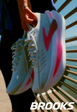 Witte sportschoenen met roze accenten en een gestructureerd bovenwerk, vastgehouden door een hand. Voorzien van veters en een gevulde zool. Merkennaam "Brooks" zichtbaar.