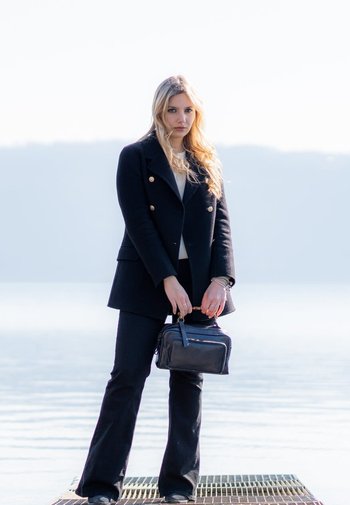 Sort dobbelbrystet frakke med guld knapper, sorte tætsiddende bukser og en rektangulær sort håndtaske. Lang bølget blond hår. Minimal baggrund.