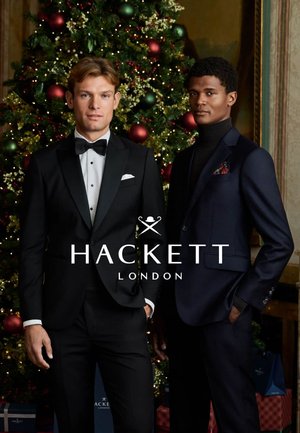 Two male models wear tailored suits. One in a black tuxedo with a bow tie, the other in a dark blue suit with a turtleneck. Christmas tree backdrop.