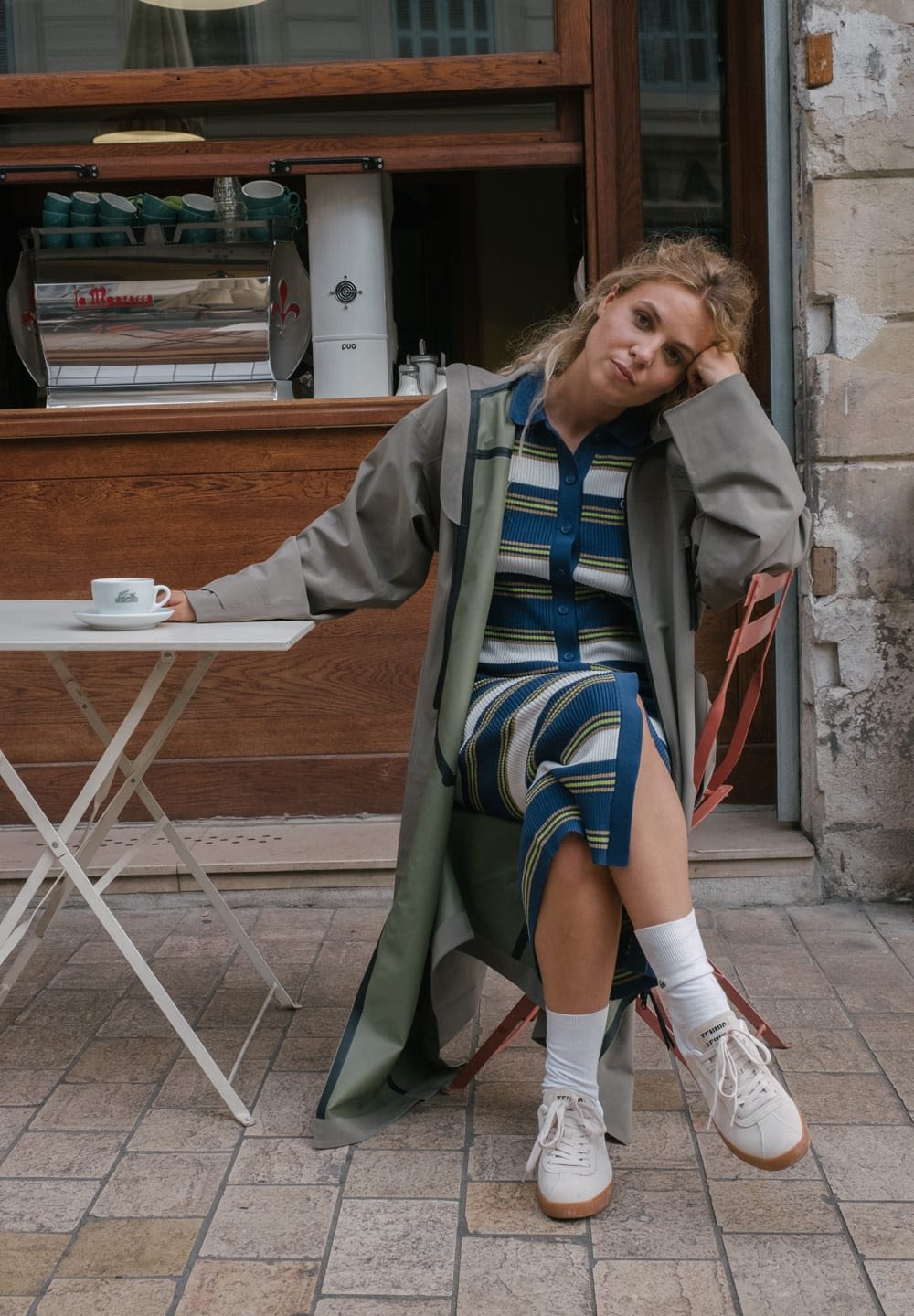 Lange grüne Oberbekleidung über einem blau gestreiften Kleid, sitzt an einem weißen Klapptisch mit einer Kaffeetasse. Trägt weiße Sneaker und Socken.