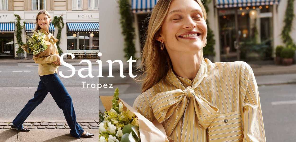 Gelb gestreifte Bluse mit übergroßer Schleife, kombiniert mit dunkelblauen Jeans mit weitem Bein. Hält einen Blumenstrauß, im Hintergrund ein Straßencafé mit gestreiftem Markisentuch.