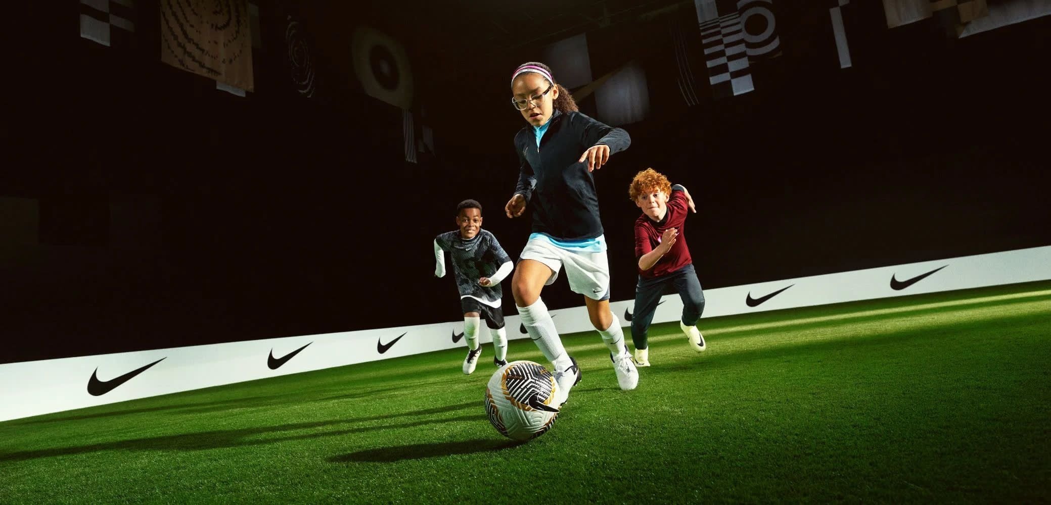 Tres niños corriendo sobre césped artificial, vestidos con ropa deportiva negra, roja y gris, y con un balón de fútbol de patrón en blanco y negro.