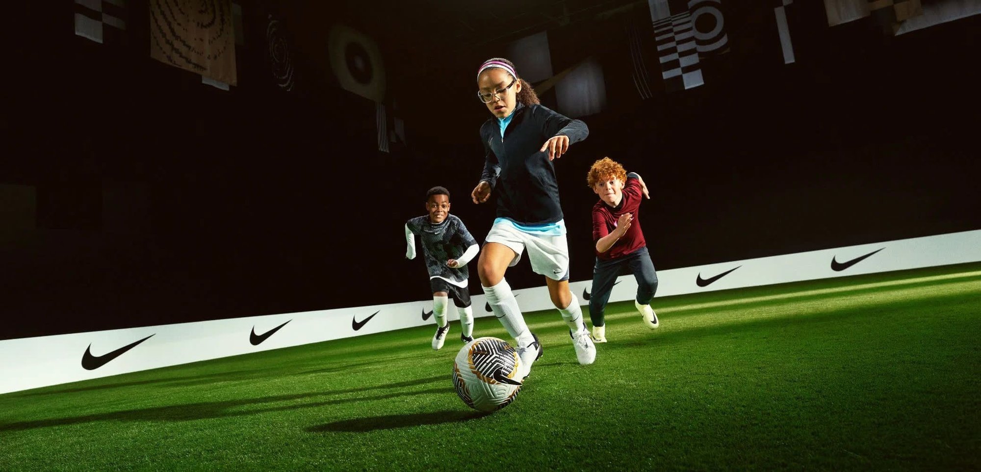 Los jugadores de fútbol juvenil corren sobre hierba sintética, vistiendo ropa atlética: chaqueta negra, camiseta azul clara, pantalones cortos blancos y un balón de fútbol colorido.