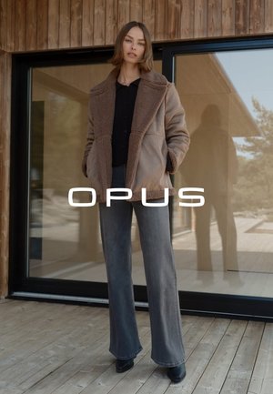 Brauner Kunstfellmantel mit breitem Kragen und seitlichen Taschen, kombiniert mit dunkelblauen, ausgestellten Jeans und schwarzen Stiefeletten. Im Glas-Hintergrund reflektiert.