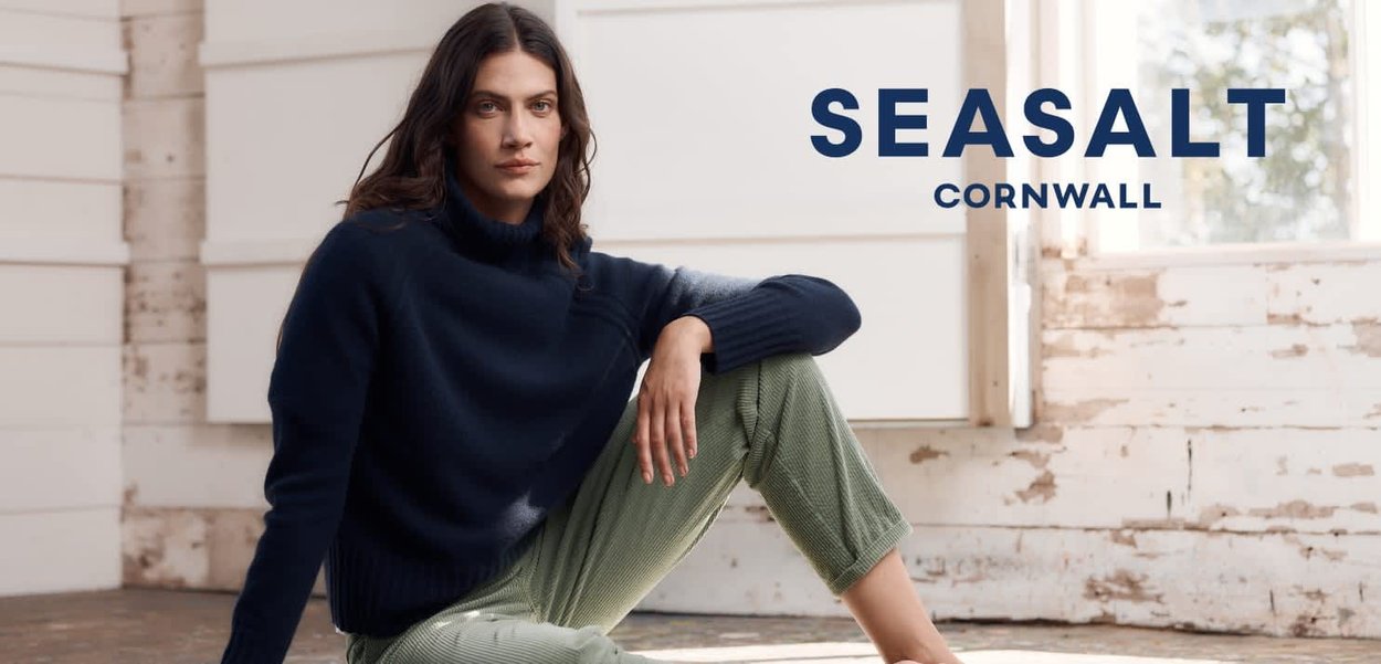 Kvindelig i en marineblå sweater og grønne bukser sidder på et trægulv i et soloplyst rustikt rum med teksten "SEASALT CORNWALL" i nærheden.