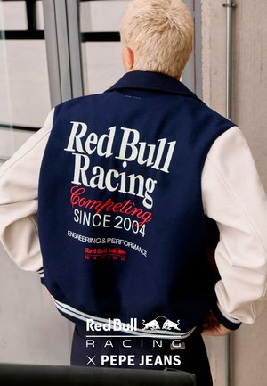 Persona con cabello rubio corto que lleva una chaqueta universitaria azul marino y blanca con el texto "Red Bull Racing Compitiendo Desde 2004" en la espalda.