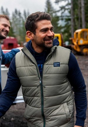 Gepolsterte olivgrüne Weste mit Reißverschluss, zwei Seitentaschen, einem hohen Kragen und einem Logo-Patch auf der Vorderseite, getragen über einem marineblauen Hoodie.