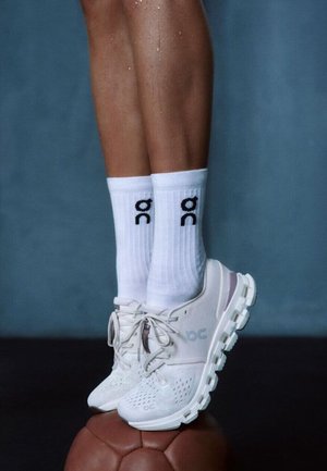 Beine mit weißen Sportschuhen und weißen Socken mit "on"-Logo, die auf einem braunen Leder-Übungsball vor blauem Hintergrund balancieren.