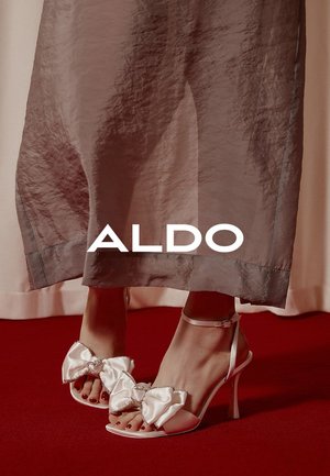 Witte satijnen sandalen met grote strikken en kristalaccenten, open teen, stilettohak, gedragen bij een vloeiende grijze jurk op een rode loper.