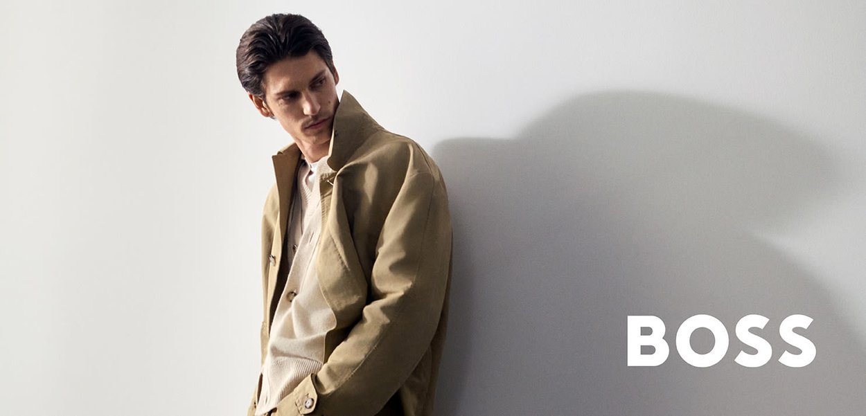 Homme en trench-coat beige et chemise claire appuyé contre un mur clair projetant une ombre, avec le mot "BOSS" en lettres blanches en gras.